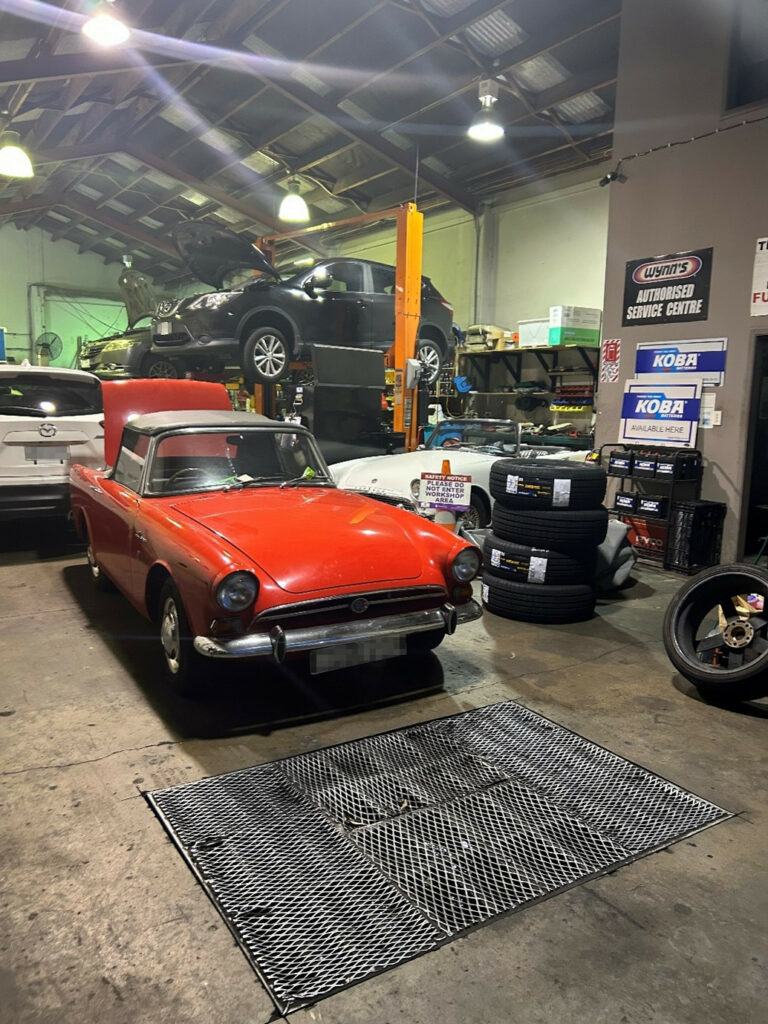Red 1967 Sunbeam Alpine parked inside an automotive workshop, with other vehicles on lifts and tyres stacked nearby.